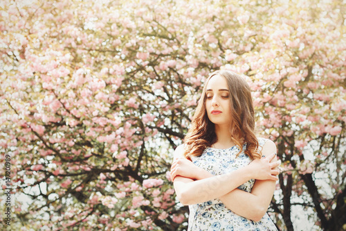 Lovely beautiful woman or girl in a dress is standing on the background of blooming cherry blossoms with sunlight, the model was seen closing her eyes