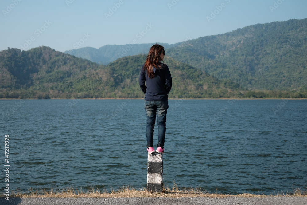 woman standing on pillar roadside front of her have big lake and mountain background. this image for nature and portrait concept