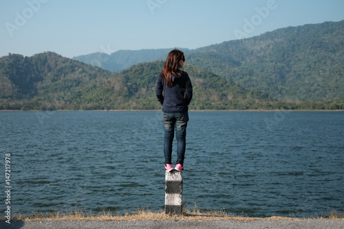 woman standing on pillar roadside front of her have big lake and mountain background. this image for nature and portrait concept