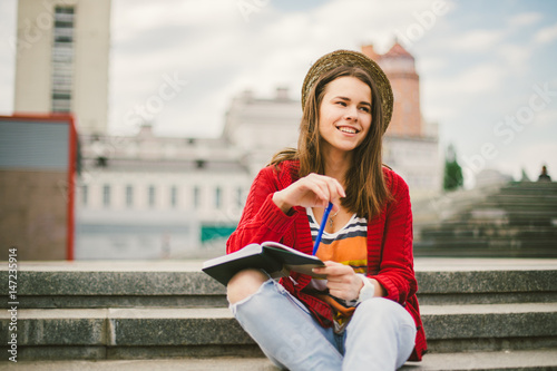 A beautiful, young Caucasian girl sitting on the street smile, the joy, sit with notebook and pen in Ruhi. In the red sweater, jeans and brown leather bag