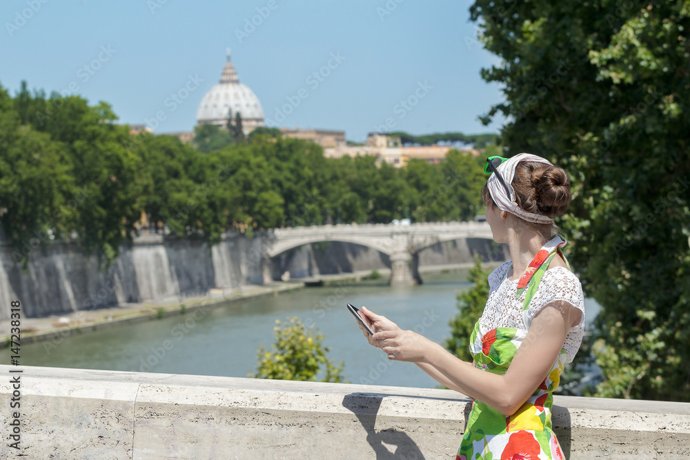 Tourist woman in flower sundress is holding tablet and looking at Rome Tiber bridge and masterpiece dome