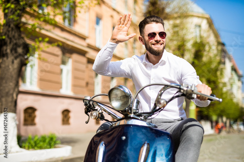 Cheerful young bearded man in helmet is sitting on scooter and say hello on city street