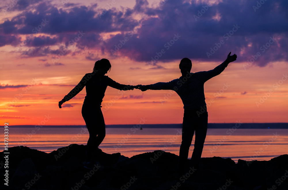 Fototapeta premium Silhouettes of woman and man on a background of colorful sunset of a cloudy day on the beach 