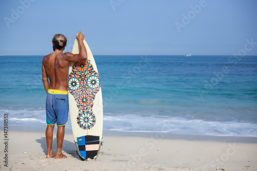 Attractive bearded curly man with a surfboard is standing with his back on a beach and looking to the ocean, located on the left side of the frame