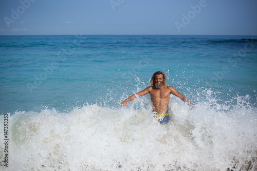 Handsome bearded curly man with naked torso and closed eyes is standing on the shore of the ocean and is opposing to the wave by his back and open arms and smiling © Dana Keli
