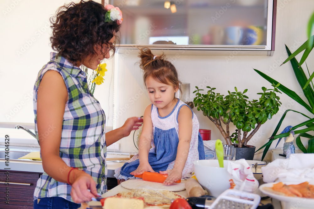 Attractive brunette female with curly hair and her cute little daughter cooking food in a home kitchen. Attractive brunette female with curly hair and her cute little daughter cooking food in a home kitchen.