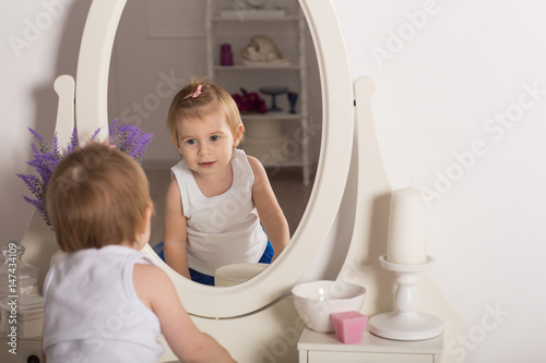 cute baby girl watching her reflection in a white bedroom with a round mirror