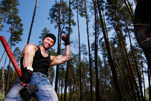 Strong healthy cheerful adult ripped man with big muscles working out with big hammer and car tyre outdoors
