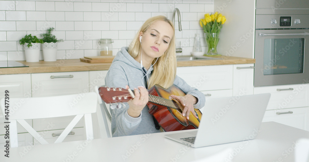 Young girl practicing guitar while sitting at home in light modern kitchen and using laptop with headphones. Young girl practicing guitar while sitting at home in light modern kitchen and using laptop with headphones.