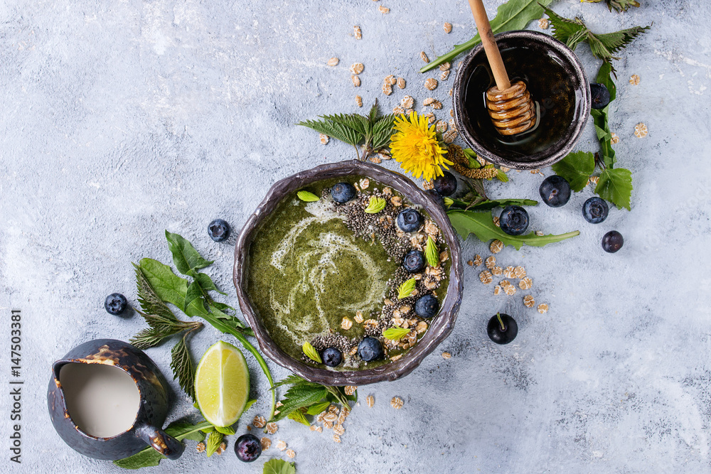 Spring green nettle and dandelion smoothie bowl served with lime, yellow flowers, young leaves, oat flakes, chia seeds, blueberries, cream and honey over gray blue texture background. Top view, space