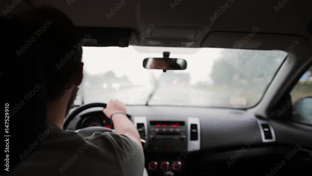 A back view of a man driving a car in the rain. Hands on a steering wheel, wipers working. A back view of a man driving a car in the rain. Hands on a steering wheel, wipers working.