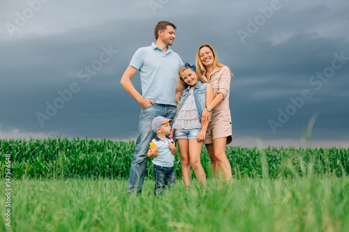 Happy family of four in the field. Thunder sky. Little boy in baseball cap. Man hugging his wife and daughter. © beatleoff