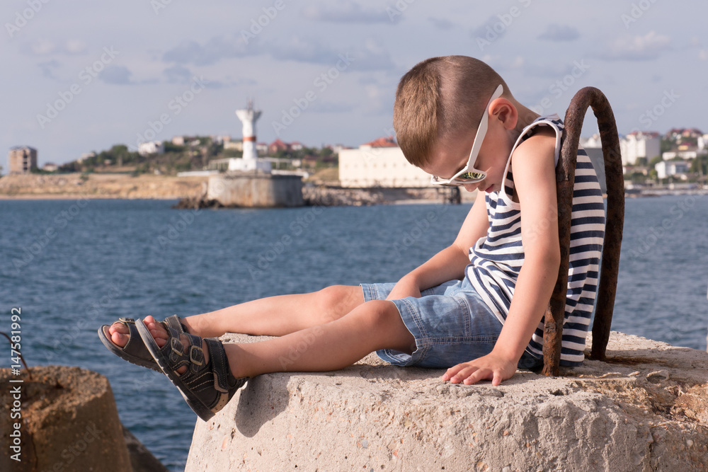 little boy in sunglasses and a vest sitting on the sea background