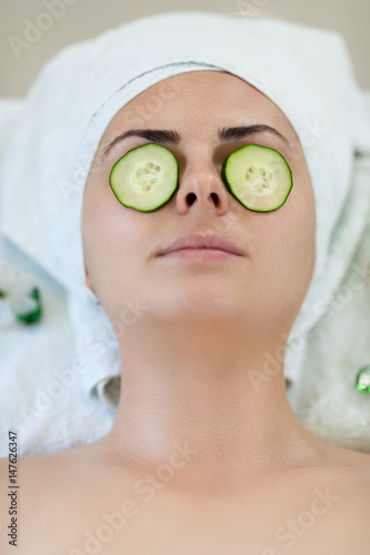 Young woman enyoing facial treatment in spa with sliced cucumbers on her eyes and towel on head. Bright scene, look from top