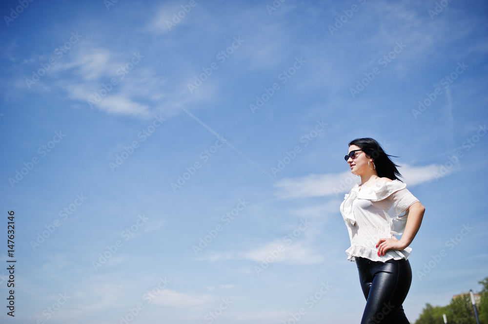 Brunette girl on women's leather pants and white blouse, sunglasses, posed against blue sky. Brunette girl on women's leather pants and white blouse, sunglasses, posed against blue sky.