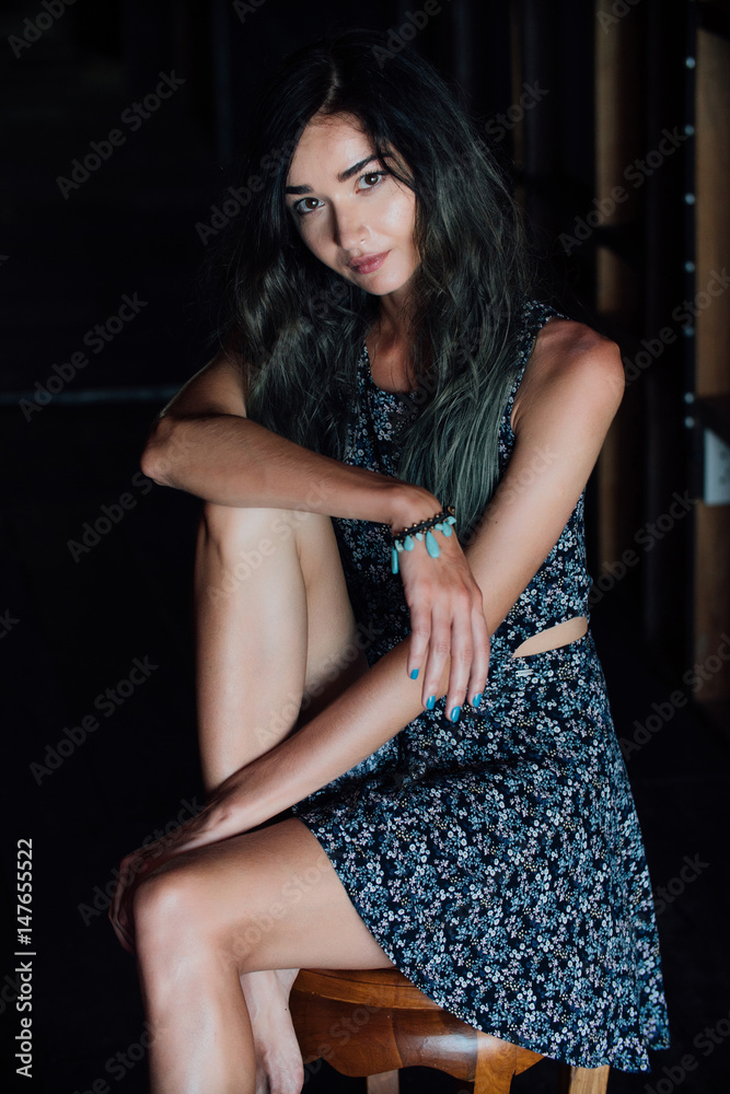 Fototapeta premium A charming young girl in a dress is sitting on a wooden chair in a cafe. Dark background.