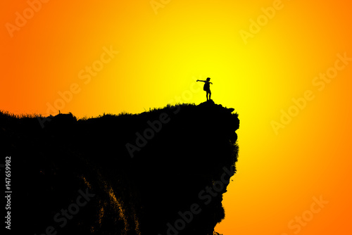 Black silhouette on orange and yellow background of woman tourist standing arms outstretched happy with success on peak mountain near the cliff at Phu Chi Fa Viewpoint, Chiang Rai, Thailand
