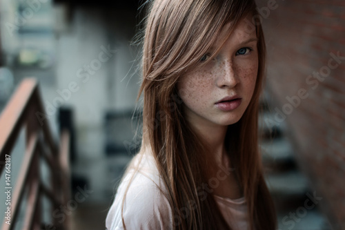 Close-up portrait of a redhead girl with freckles and blue eyes standing on the stairs and looking at camera