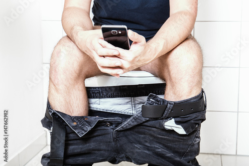 Young man sittinging on the toilet with smartphone in his hands photo