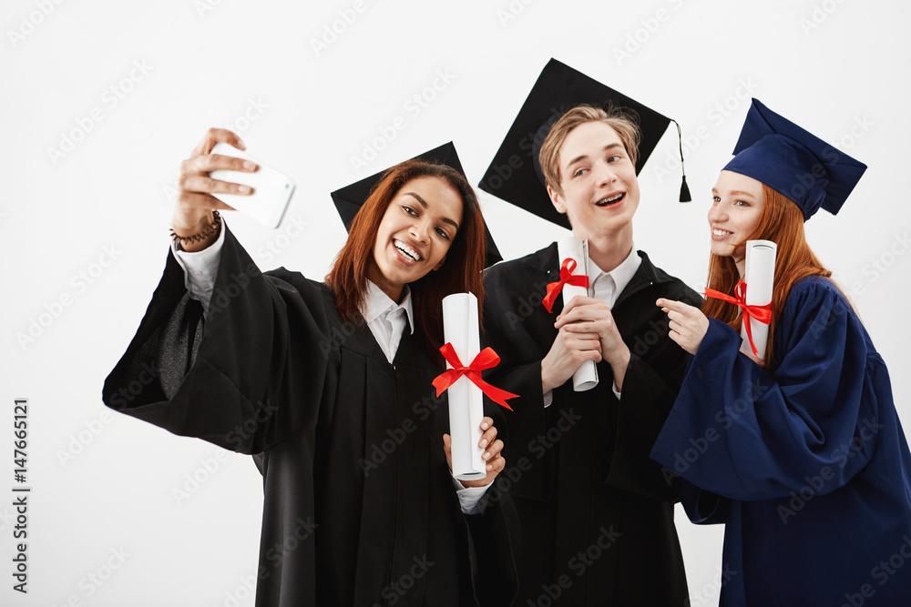 Three international graduate friends rejoicing in mantles making a selfie on a phone. Future lawyers or doctors or engineers having fun with diplomas. White background. Three international graduate friends rejoicing in mantles making a selfie on a phone. Future lawyers or doctors or engineers having fun with diplomas. White background.
