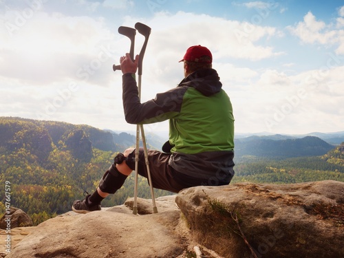 Fotografija Tired tourist with medicine crutch  and broken leg fixed in immobilizer resting on summit
