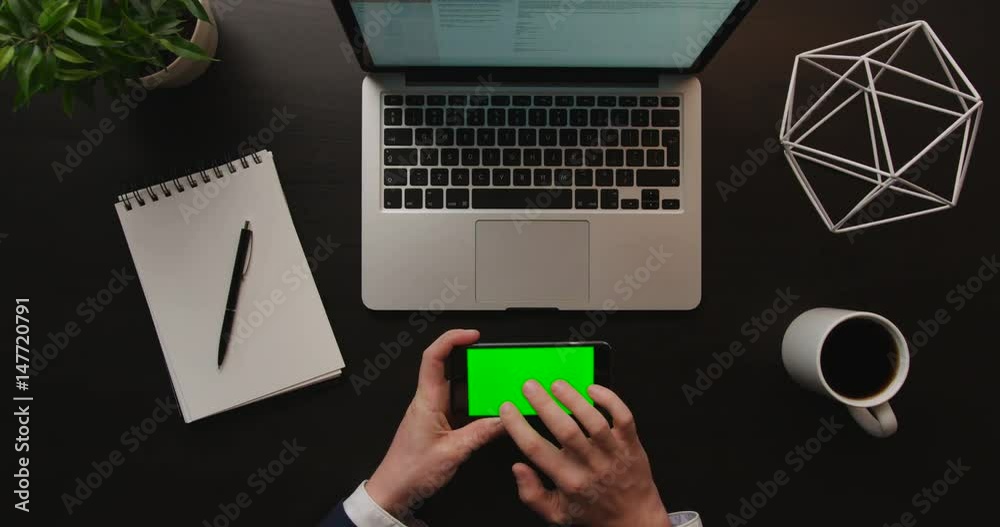 Business man using smart phone, checking information on cell phone with green screen on brown background with laptop. Office desk background. Slow motion. Top view. Chroma key. Horizontal position. Business man using smart phone, checking information on cell phone with green screen on brown background with laptop. Office desk background. Slow motion. Top view. Chroma key. Horizontal position.
