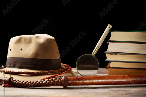 Fedora hat with bullwhip near magnifying glass and old books on black background. Adventure concept