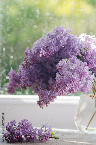 Beautiful lilac flowers in a clear glass vase on the table in front of the window, selective focus
