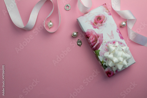 Pink background with wrapped gift, white satin ribbon and pearl and crystal charms for Mothers day or birthday