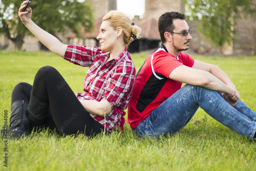 Guy and girl back-to-back sitting in grass, neglecting each other. She is taking selfie