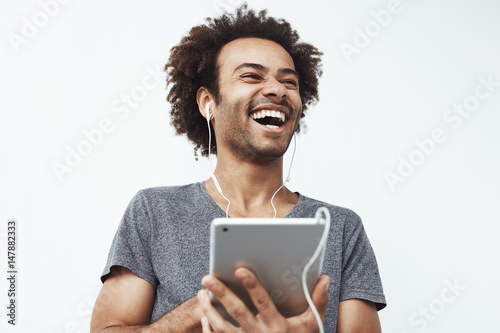 Portrait of cheerful happy african man in headphones laughing holding tablet talking or watching and enjoying a comedy show or browsing over white background Fototapet