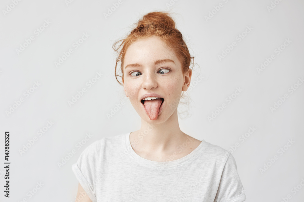 Portrait of crazy and silly yet beautiful freckled girl with eyes crossed, puting out toungue fooling around. Pretending being at audiologist. Easy lifestyle concept on white. Portrait of crazy and silly yet beautiful freckled girl with eyes crossed, puting out toungue fooling around. Pretending being at audiologist. Easy lifestyle concept on white.