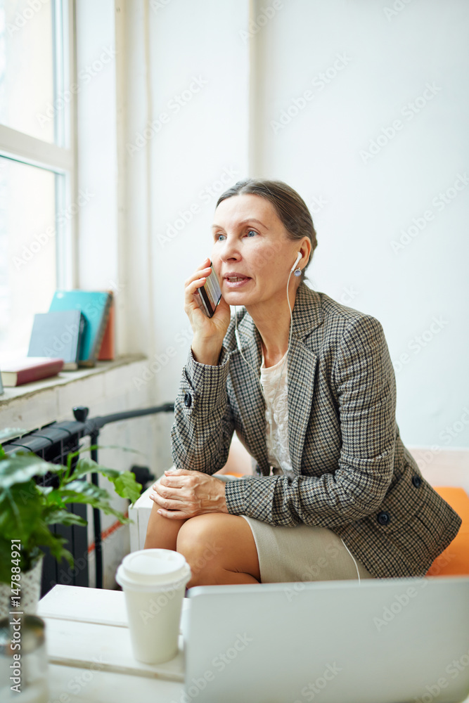 Busy senior entrepreneur sitting in spacious office lobby with paper cup of coffee and discussing project details on smartphone, portrait shot Busy senior entrepreneur sitting in spacious office lobby with paper cup of coffee and discussing project details on smartphone, portrait shot