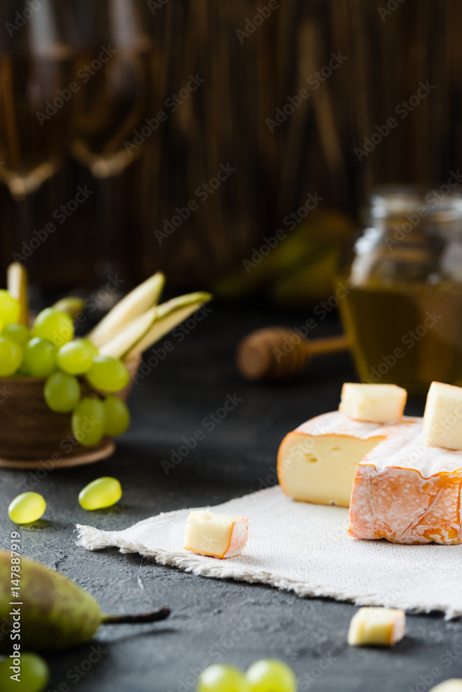 French soft cheese from Brittany region and sliced brie with pear, green grapes, honey and glasses of white wine on dark rustic background French soft cheese from Brittany region and sliced brie with pear, green grapes, honey and glasses of white wine on dark rustic background