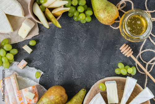 Platter of French soft cheeses from Normandy and Brittany regions, and brie sliced with honey, pear and green grapes on dark rustic background with copy space, top view