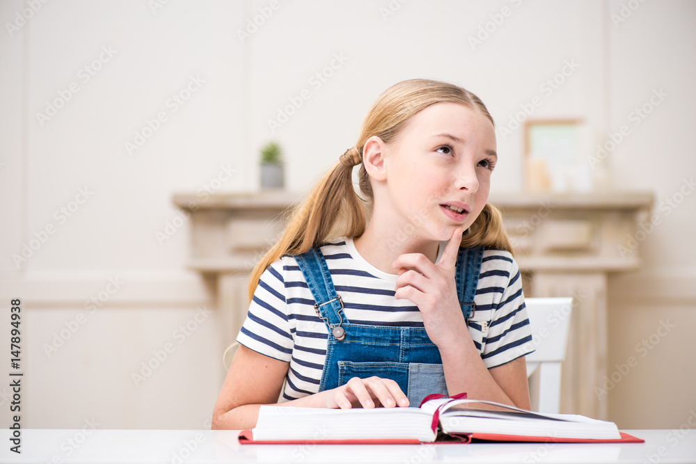 Cute little girl sitting at table with book and thoughtfully looking away Cute little girl sitting at table with book and thoughtfully looking away