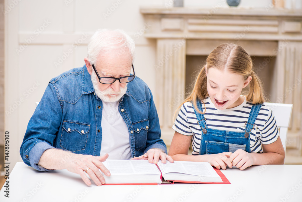 Smiling girl and grandfather in eyeglasses reading book together at home Smiling girl and grandfather in eyeglasses reading book together at home