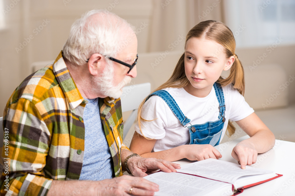 Grandfather and granddaughter sitting at table and reading book together Grandfather and granddaughter sitting at table and reading book together