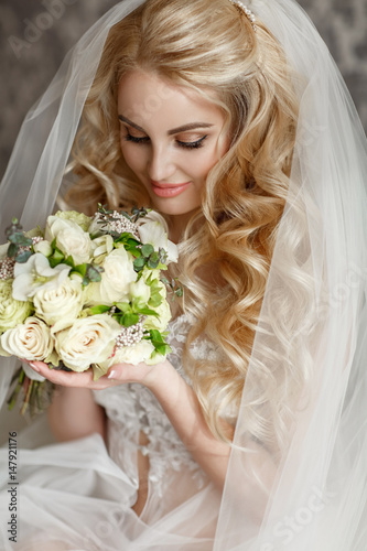 Beautiful young bride at home wearing fashion wedding dress. Newlywed woman cozy home interior. Happy gilr bride in love waiting for groom and holding bridal flowers bouquet