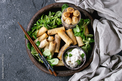 Fried spring rolls with white yogurt sauces, served in terracotta plate and fry basket with fresh green salad and chopsticks over black texture background. Flat lay, space. Asian food