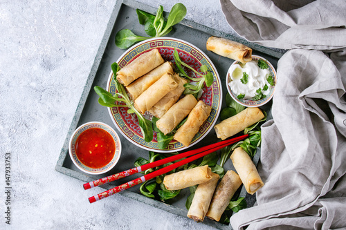 Fried spring rolls with red and white sauces, served in china plate on wood tray with fresh green salad and wooden chopsticks over gray blue texture background. Flat lay, space. Asian food