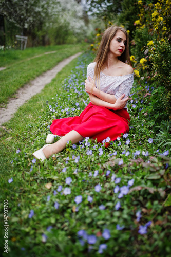 Portrait of sitiing beautiful girl with red lips at spring blossom garden on grass with flowers, wear on red dress and white blouse. © AS Photo Family