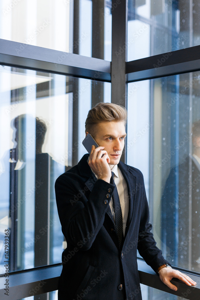 Concentrated young entrepreneur in stylish suit talking to his client on smartphone while standing by panoramic window, portrait shot Concentrated young entrepreneur in stylish suit talking to his client on smartphone while standing by panoramic window, portrait shot