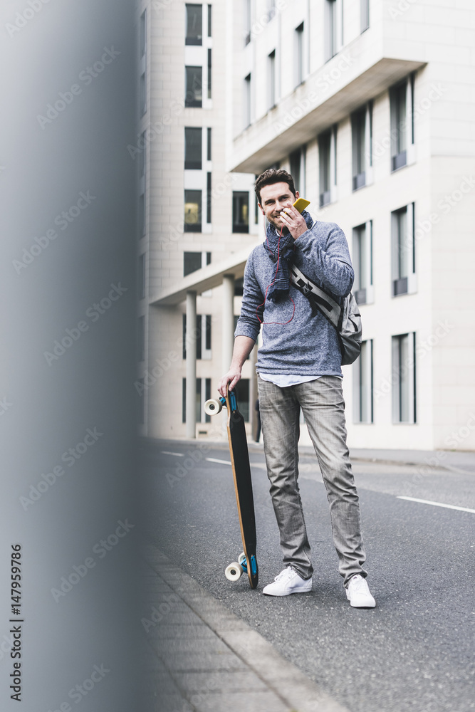 Businessman carrying skateboard, using smartphone and earphones Businessman carrying skateboard, using smartphone and earphones