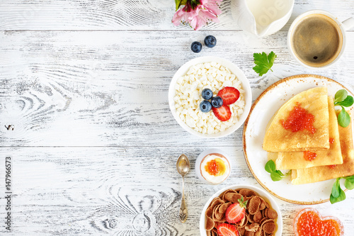 Overhead of traditional russian breakfast on white wooden table with copy space. Pancakes with caviar, cottage cheese with strawberry and blueberry, cornflakes, boiled egg and coffee.