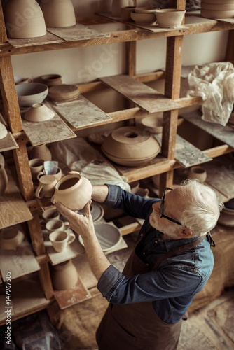 Senior potter in apron and eyeglasses examining ceramic bowl at workshop