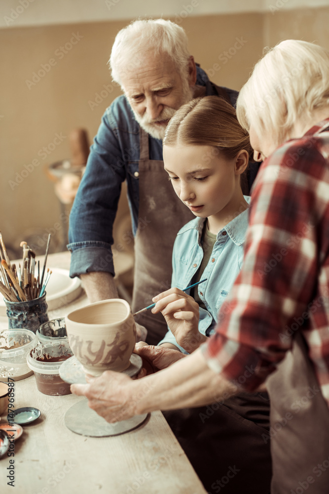Side view of girl painting clay pot and grandparents helping at workshop Side view of girl painting clay pot and grandparents helping at workshop