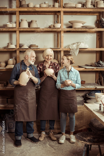 Granddaughter and grandparents standing and holding clay vase and bowls against wall with pottery goods