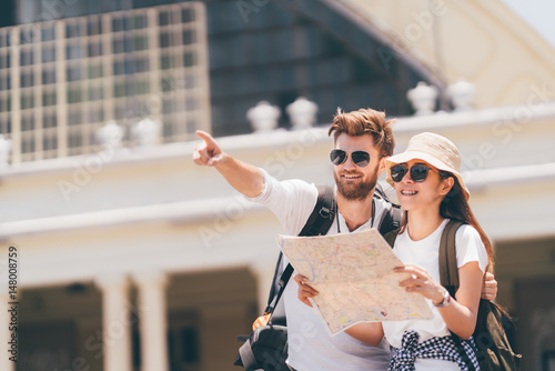 Multi-ethnic traveler couple using local map together on sunny day, man pointing forward to copy space. Honeymoon trip, backpacker tourist, Asia city tourism, or summer holiday vacation travel concept