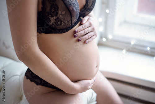 Closeup beautiful pregnant lady in elegant pose in black bra and panties. Portrait of sexy fashion model girl in lace lingerie. Beauty woman with attractive body indoors. Close-up female naked belly